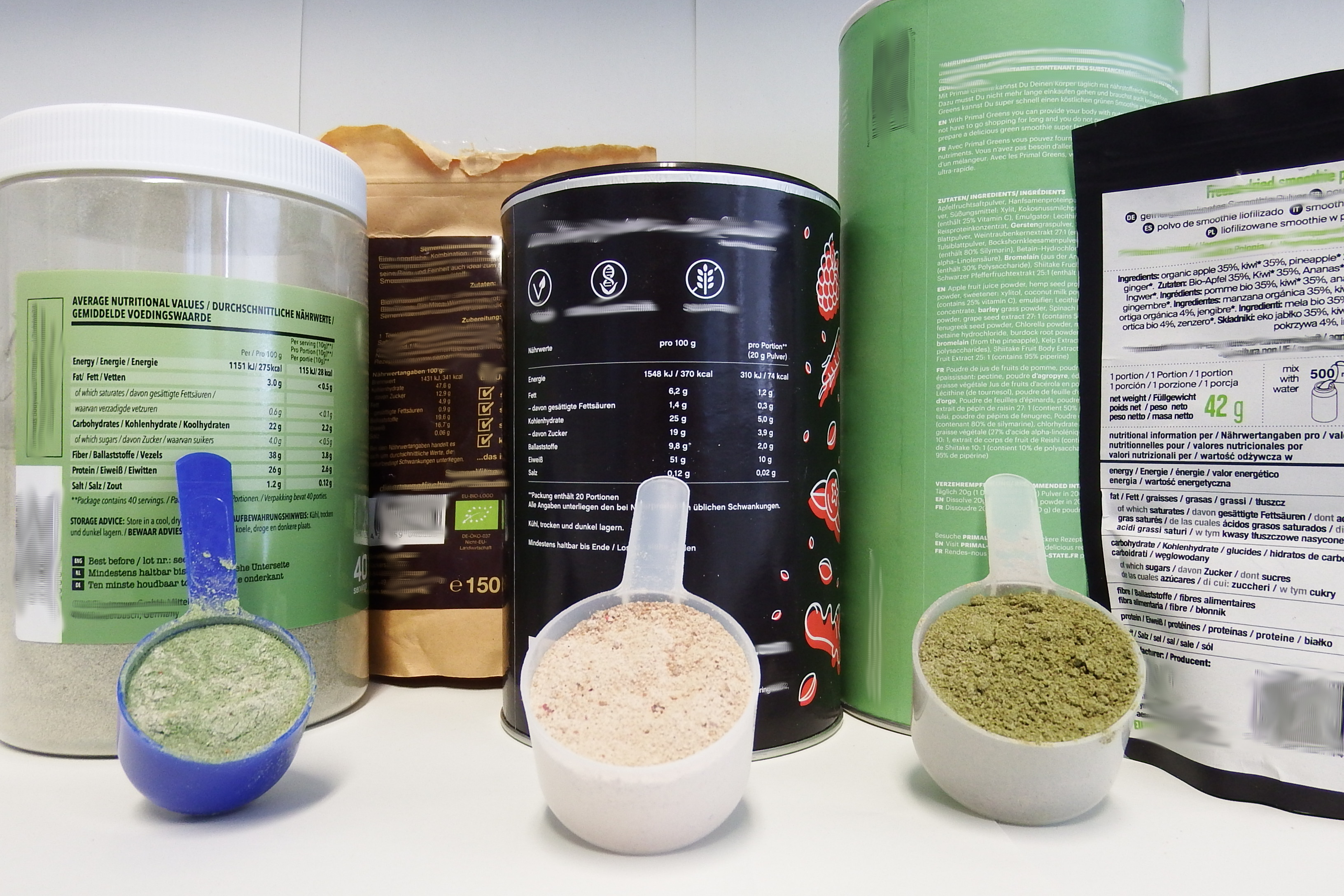 Das Bild zeigt fünf verschiedene Verpackungen von Smoothiepulvern. Davor sind drei Dosierlöffel mit Smoothiepulvern in grüner und hellroter Farbe abgebildet.